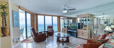 A spacious living room with a large window overlooking the ocean, a ceiling fan, and a cozy seating area with a sofa and chairs.