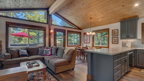 A spacious living room with a large sofa, a dining area with wooden chairs, a kitchen with grey cabinets, and large windows offering a view of trees.