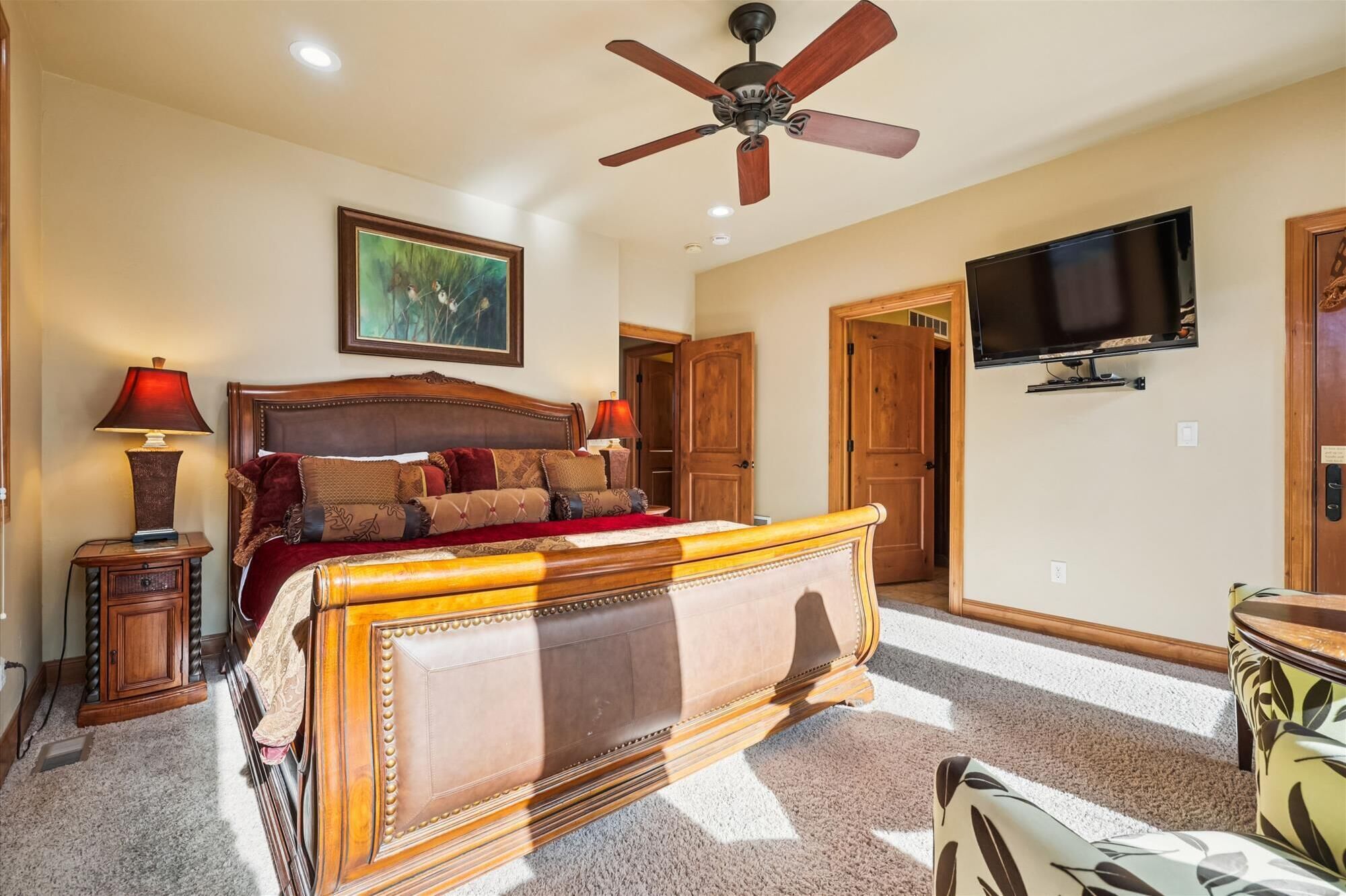 Un dormitorio con una cama grande, un ventilador de techo, un televisor montado en la pared y una puerta que da a otra habitación.