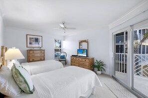 Une chambre avec deux lits, un ventilateur de plafond, une télévision, une commode en bois, une chaise, une lampe et une grande fenêtre donnant sur un beau paysage.