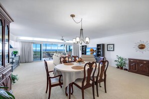 Un vaste salon comprenant un coin repas, de grandes fenêtres, un lustre et une vue sur l’océan.