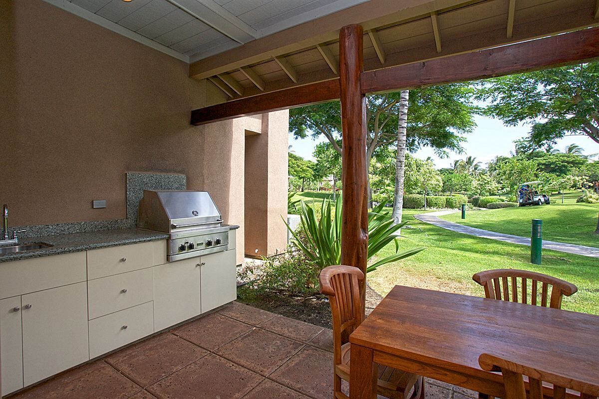 An outdoor kitchen area with a grill, a table, and chairs, surrounded by a patio with a view of a well-maintained lawn and trees.