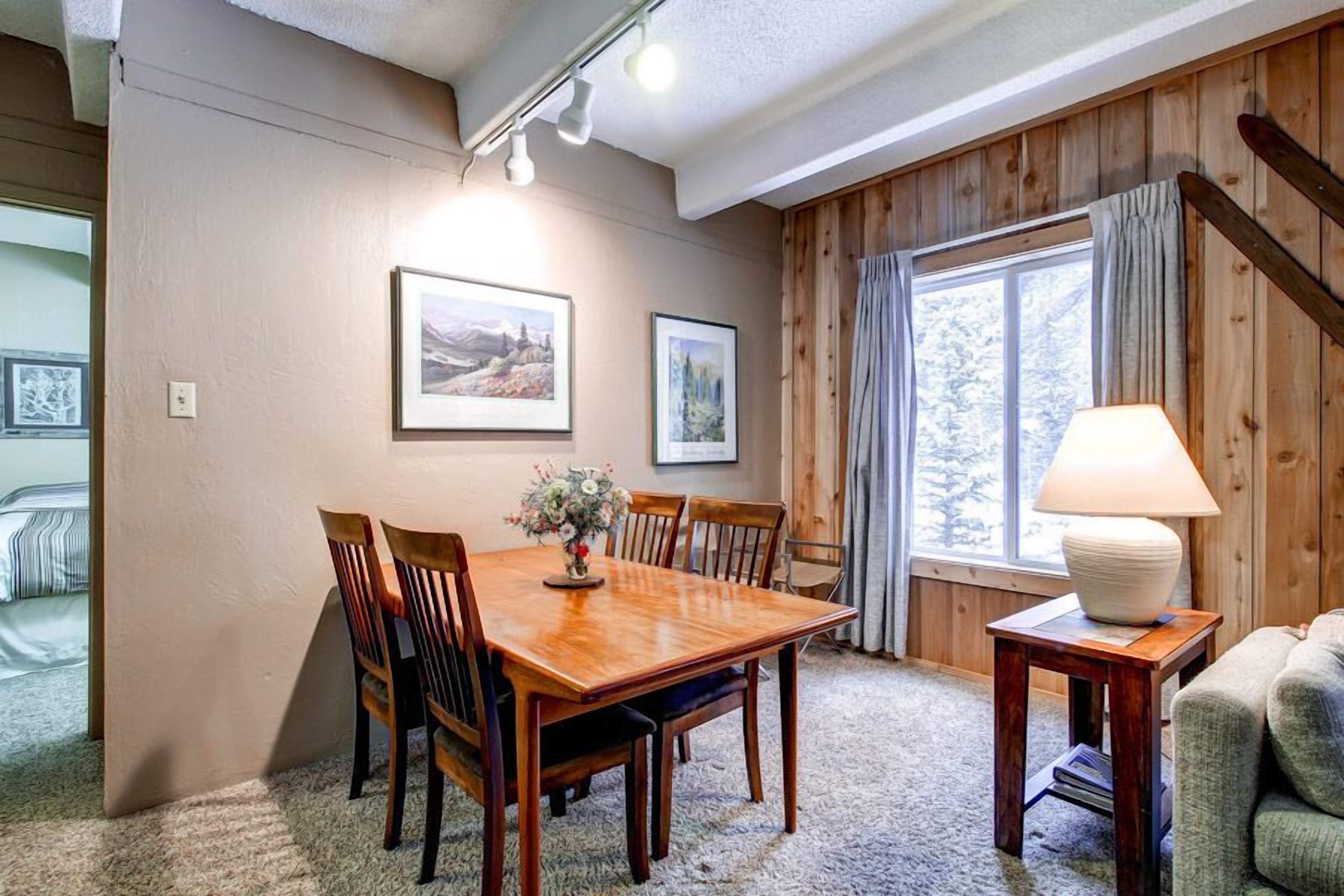 A cozy room with a wooden dining table, chairs, and a lamp. There are framed pictures on the wall and a window with curtains.