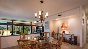 A dining area with a round glass table, wicker chairs, a chandelier, and a view of the outdoors through large windows. - Kaanapali Royal L-101  Located on Golf Course (Lahaina)