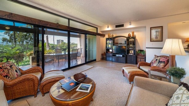 A spacious living room with a large sliding glass door leading to a balcony, a television, and a cozy seating area with wicker furniture. - Kaanapali Royal L-101  Located on Golf Course (Lahaina)