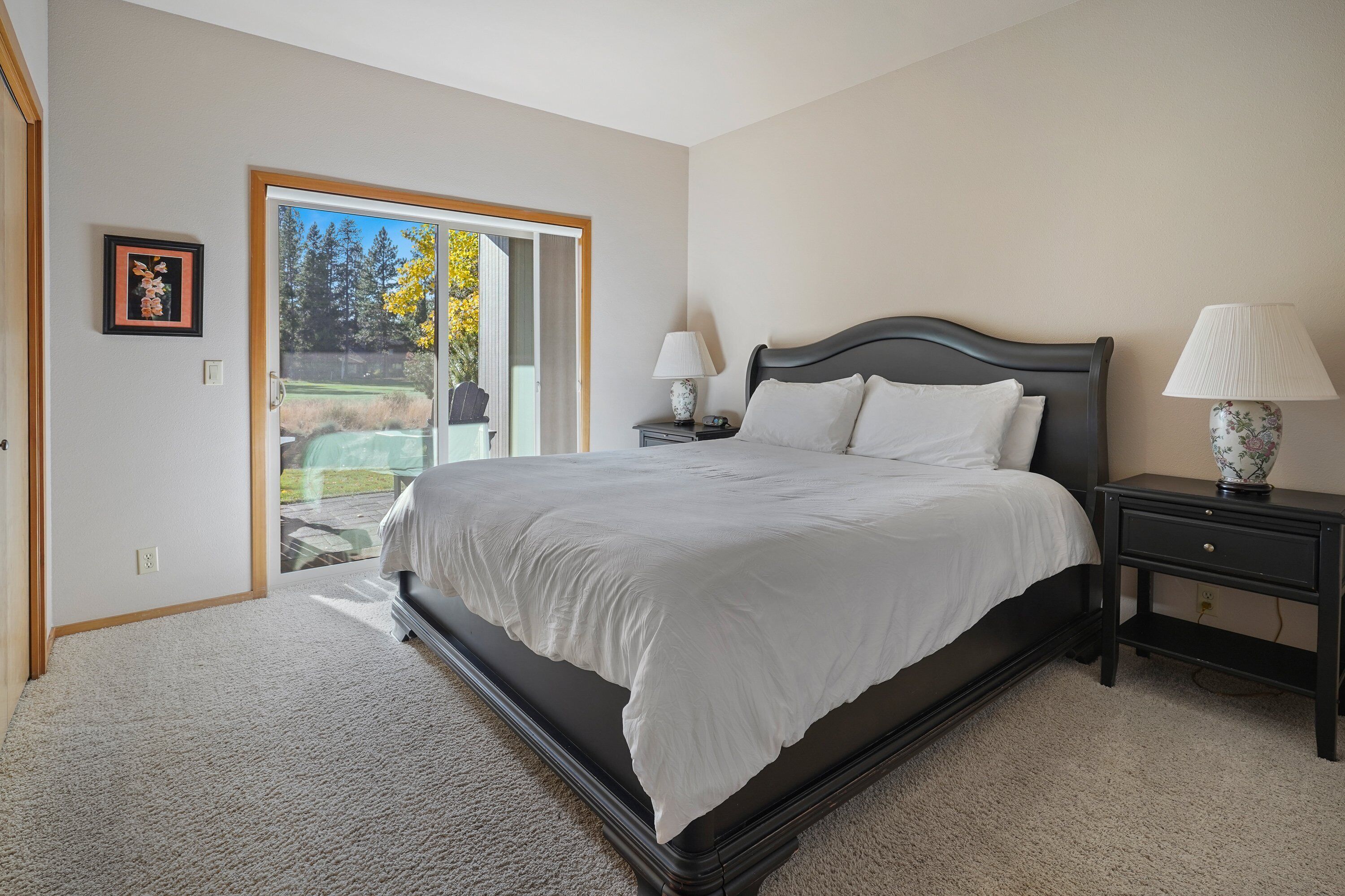 A bedroom with a large bed, two bedside tables with lamps, a framed picture on the wall, and a sliding glass door leading to a patio.