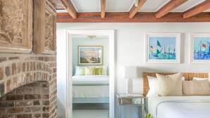 A bedroom with a brick fireplace, a bed with white bedding, and two framed paintings on the wall. - Entire 1842 Downtown Home with Private Courtyard + Parking by Lucky Savannah (Savannah)
