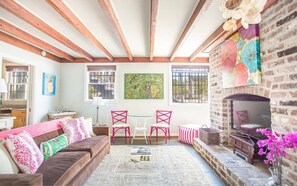Un salon chaleureux avec une cheminée, des poutres en bois, un canapé, une table basse et deux fauteuils roses.