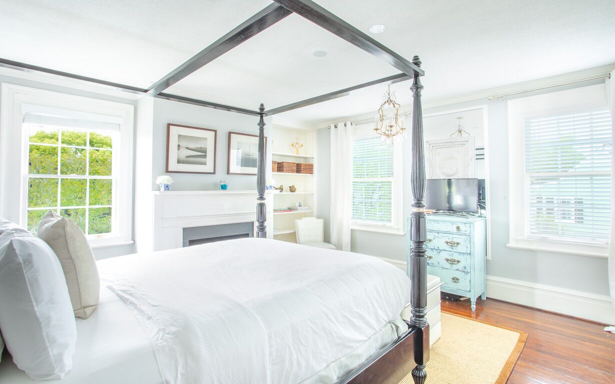 A four-poster bed in a bright room with large windows, a dresser, and a chandelier.