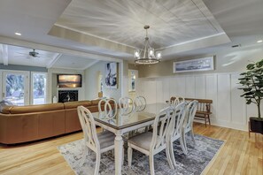 A dining area with a glass-top table and white chairs, a brown leather sofa, and a chandelier. - 52 Gloucester-The Perfect Pool and 8 minute bike ride to Shipyard Beach Club (Hilton Head Island)