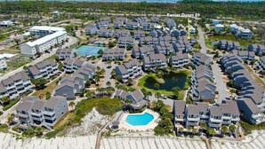 Vista aérea de una zona residencial con varias casas de dos pisos, una piscina central y una playa con un paseo de madera.