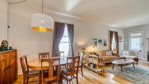 Un salon spacieux avec une table à manger en bois, un canapé et une table basse. Une grande fenêtre, ornée de rideaux, laisse entrer la lumière, et un luminaire suspendu éclaire la pièce.