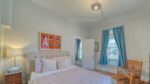 Une chambre à coucher comprenant un lit, une table de chevet, une lampe, une chaise, une porte et une fenêtre avec des rideaux.