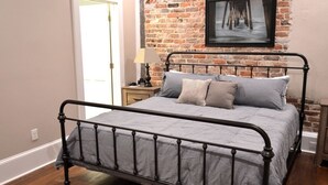 A bedroom with a metal bed frame, a brick wall, a framed picture, and a nightstand with a lamp.
