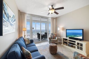 A living room with a sofa, armchair, ottoman, TV, ceiling fan, and a balcony with a view of the ocean.