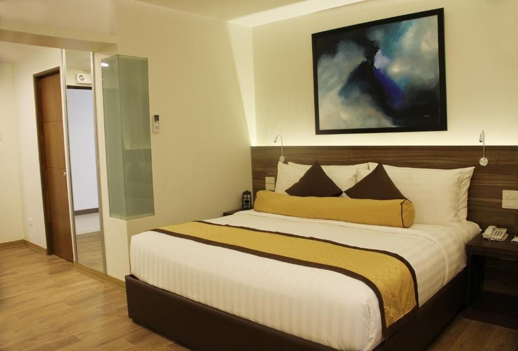 A hotel room with a large bed, a painting on the wall, a bedside table with a phone, and a door leading to another room.