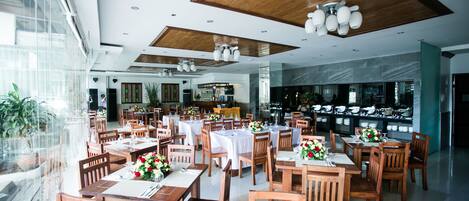 A spacious restaurant with wooden tables and chairs, white tablecloths, and a modern ceiling design.