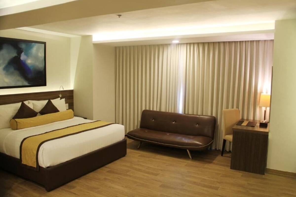 Habitación de hotel con una cama grande, un sofá de cuero marrón, un escritorio pequeño y un ventanal con cortinas.