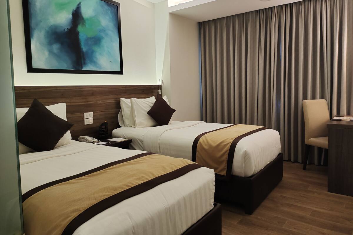 Habitación de hotel con dos camas, un escritorio, una silla, un cuadro en la pared y una ventana con cortinas.