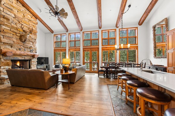 A spacious living area with a stone fireplace, wooden beams, and large windows offering views of the outdoors.
