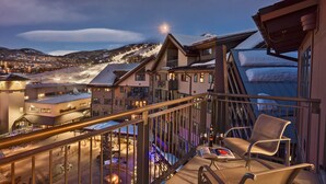 A balcony with a chair and a table, overlooking a snow-covered ski slope and a town at dusk.