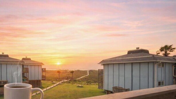 A sunset view from a balcony overlooking two buildings with a wooden deck in the foreground. - Ocean View Escape | Balcony Views + Boardwalk to the Beach (Port Aransas)