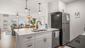 A modern kitchen with white cabinetry, a stainless steel refrigerator, and pendant lighting. - Ocean View Escape | Balcony Views + Boardwalk to the Beach (Port Aransas)