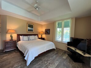 A bedroom with a bed, two bedside tables with lamps, a ceiling fan, a window with blinds, and a TV on a stand.