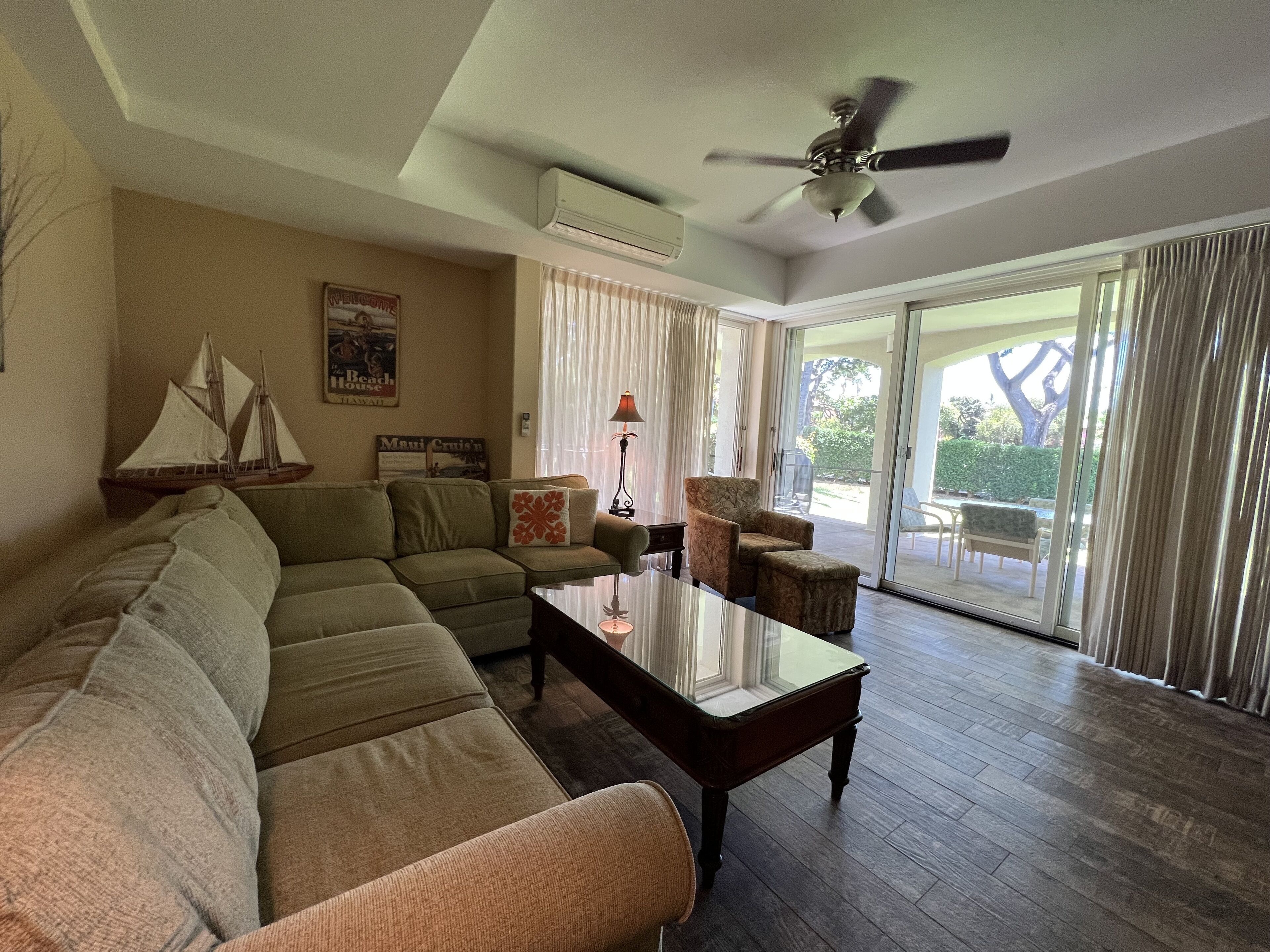 A living room with a sectional sofa, a glass coffee table, a patterned armchair, and a ceiling fan. There is a window with curtains and a view of the outdoors.