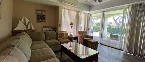 A living room with a sectional sofa, a glass coffee table, a patterned armchair, and a ceiling fan. There is a window with curtains and a view of the outdoors.