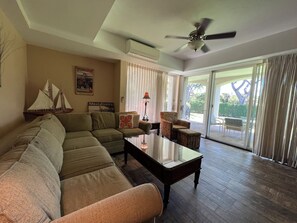 A living room with a sectional sofa, a glass coffee table, a patterned armchair, and a ceiling fan. There is a window with curtains and a view of the outdoors.