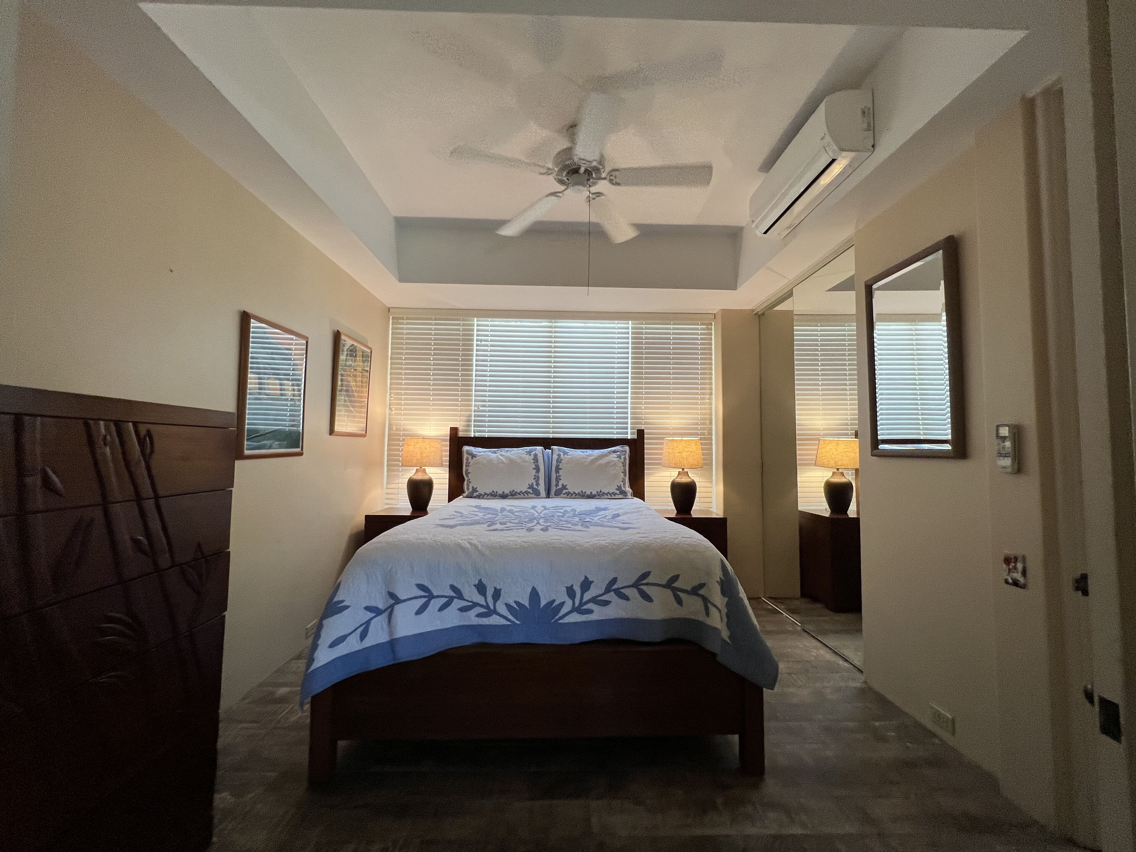 A bedroom with a bed, two bedside tables with lamps, a ceiling fan, and a large window with blinds.