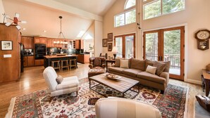 A spacious living area with a large sofa, a coffee table, and a patterned rug. There is a kitchen in the background with wooden cabinets and a dining area with a table and chairs. - Big Springs by Tahoe Getaways - Luxury Northstar 6 BR - Free Ski Shuttle to Slopes (Truckee)