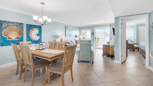 Een ruime woonkamer met een eettafel, stoelen en een kroonluchter. Er zijn kunstwerken aan de muur en uitzicht op het buitenland.