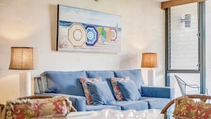 Ein modernes Wohnzimmer mit einem blauen Sofa, floralen Kissen, einem Strandbild und einem Glastisch.