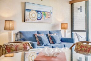 Un soggiorno moderno con un divano blu, cuscini a motivi floreali, un quadro a tema spiaggia e un tavolino da caffè in vetro.