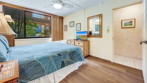 Ein Schlafzimmer mit einem Bett, einem Deckenventilator, einer Kommode, einem Fernseher und einem Fenster mit Blick auf Palmen.