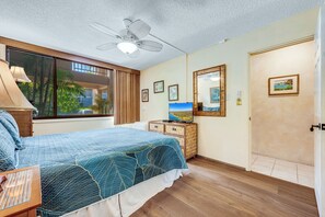 A bedroom with a bed, a ceiling fan, a dresser, a TV, and a window with a view of palm trees.