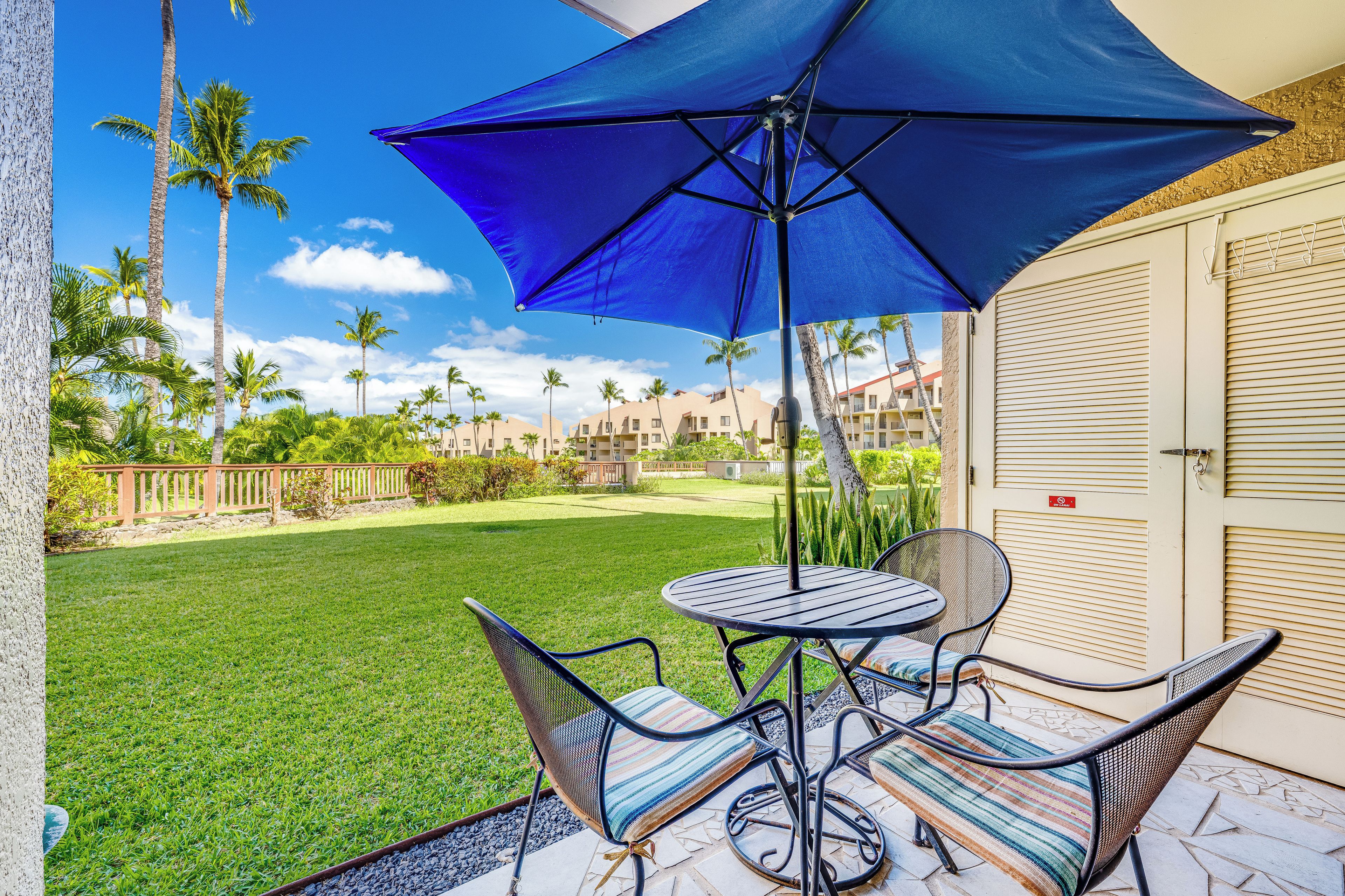 A patio with a table and chairs, a blue umbrella, and a view of a lawn and buildings.