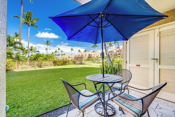 A patio with a table and chairs, a blue umbrella, and a view of a lawn and buildings.