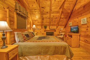 A log cabin bedroom with a bed, two bedside lamps, a television, a fireplace, and wooden walls. - Pigeon Forge | Pool Table | Hot Tub | WiFi | Resort Pool | Fireplace (PIGEON FORGE)