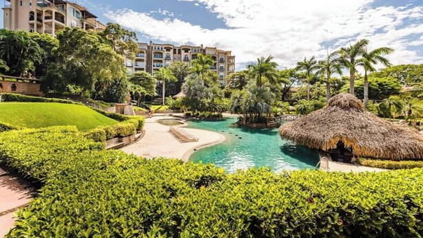 Un complexe hôtelier doté d’une piscine, d’une végétation luxuriante et d’une structure à toit de chaume.