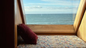 Una habitación pequeña y triangular con suelo de madera y una alfombra colorida con estampado. Un cojín rojo está apoyado contra la pared, y la habitación tiene vistas al océano a través de un ventanal grande.