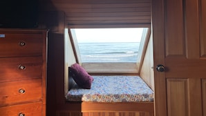 Una habitación pequeña y cerrada con techo inclinado, un aparador de madera, una cama individual con colchón estampado y vista al océano a través de un tragaluz.