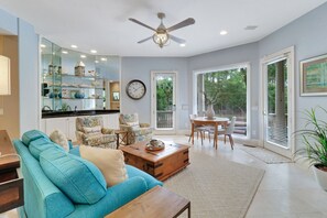 Un salon spacieux avec un canapé de couleur turquoise, une table basse en bois et un coin repas comprenant une table et des chaises en bois. On y trouve également un grand miroir, une horloge et un ventilateur de plafond.