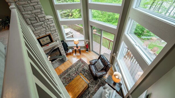 A spacious living area with a stone fireplace, large windows, and a view of a wooden deck and greenery. - Hot Tub, Fire Pit & Stunning Lake Views (Oakland)