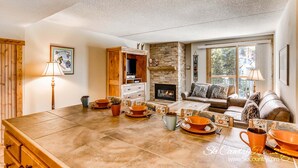 A cozy living room with a fireplace, a dining area with orange tableware, and a sofa with patterned cushions. - Steps to Adventure, Remodeled Kitchen, Wi-Fi, Ski in/out, Gas Fireplace TE411 (Breckenridge)