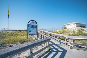 A wooden boardwalk leading to a beach area with a safety sign, a dune access point, and a beach house in the background.