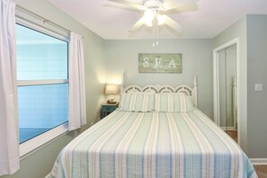 A bedroom with a bed, a ceiling fan, a window with curtains, a nightstand with a lamp, and a wall with the word 'SEA' painted on it.