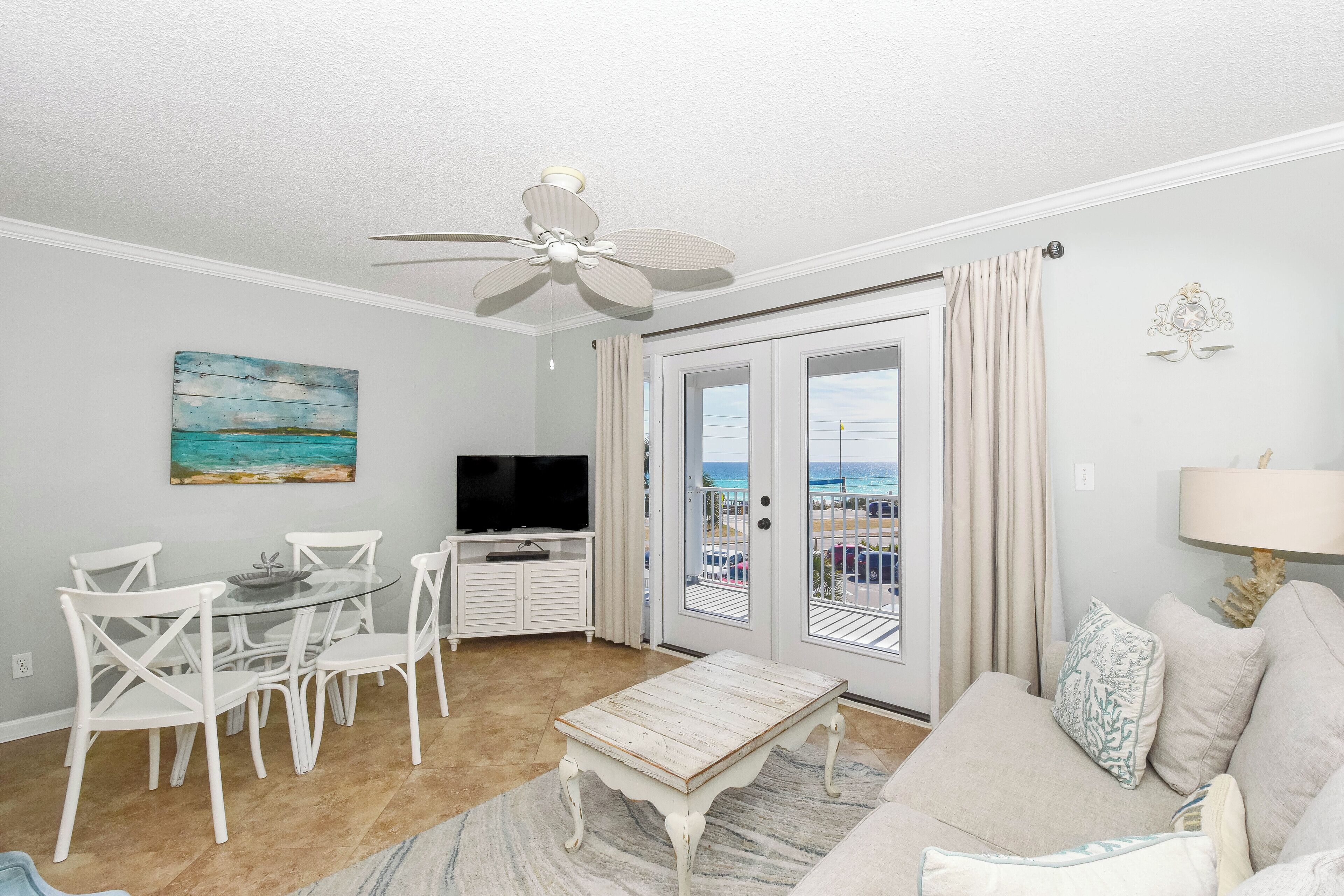 A living room with a ceiling fan, a television, a dining table with chairs, a sofa, and a view of the beach through the glass doors.